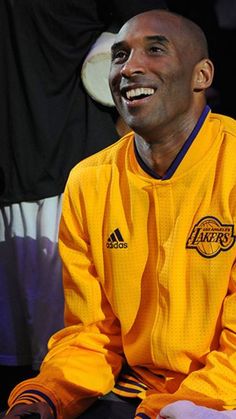 a smiling man in a lakers jersey sits on the sidelines at a basketball game