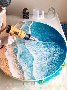 a woman is using a drill to paint a beach scene on a plate with sand and water