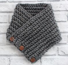 a gray knitted neck warmer with buttons on the front and side, against a white brick wall