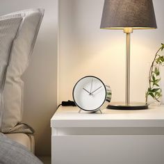 an alarm clock sitting on top of a white nightstand next to a lamp and pillow