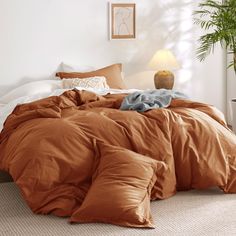 a bed with an orange comforter and pillows in a white room next to a potted plant