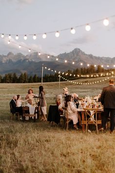 a group of people sitting around a table in the middle of a field with string lights