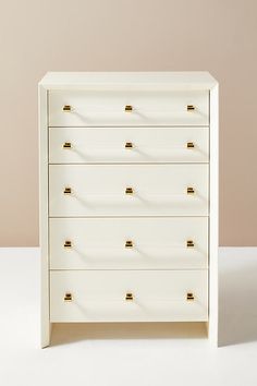 a white chest of drawers with gold handles and knobs on the bottom, against a beige wall