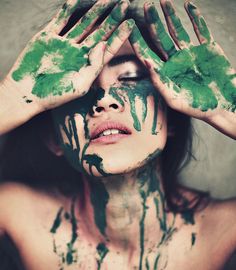 a woman with green paint on her face holding her hands up to her head and covering her eyes