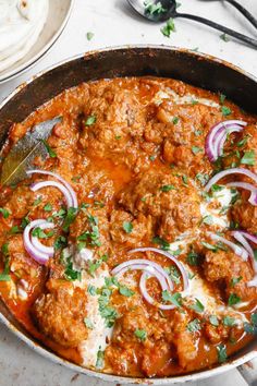 a skillet filled with meat and onions on top of a white table next to silver utensils