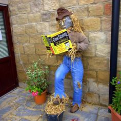 a scarecrow reading a book in front of a brick wall with potted plants