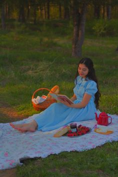 a woman sitting on a blanket in the grass reading a book next to a picnic basket