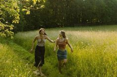 two young women walking through tall grass in the woods, one is holding her hand