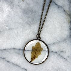 This tiny spring oak leaf was hand gathered in early spring and pressed to preserve it's beauty. It has been encased in jewelry grade resin with an antique bronze plated circle bezel. The matching chain is 24 inches in length. Chain is finished with a lobster claw clasp. Necklaces will arrive attached to card shown in photos- Ready for gifting! Want another type of leaf? Check out our other listings! Use this link: goo.gl/kaJUsJ Other Options Include: Maple Red Maple Silver Maple White Oak Red O Nature-inspired Leaf-shaped Jewelry With Pressed Flowers, Nature-inspired Jewelry With Natural Inclusions In Round Pendant, Leaf Shaped Pressed Flowers Jewelry Gift, Leaf-shaped Pressed Flowers Jewelry Gift, White Oak Leaf, Oak Leaf Necklace, Spring Leaves, Real Leaf, Terrarium Necklace