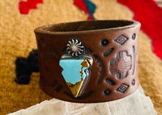 A one of a kind 1.5 inch hand stamped and stained leather cuff in a dark brown, with a mixed southwestern stamping pattern.  The cuff has an artisan handmade sterling silver piece that has a stamped symbol of the sun on the top and an arrow  with a beautiful piece of Baja turquoise in the center that measures 1.25 inches in length and .75 at the widest point.  All of our bracelets are hand crafted by us from the finest leather and constructed to last.  We take our time researching pieces for our Rustic Brown Cuff Bracelet With Concho, Western Style Brown Cuff Bracelet As Gift, Artisan Brown Cuff Bracelet With Concho, Southwestern Hand Tooled Brown Cuff Bracelet, Artisan Brown Concho Cuff Bracelet, Southwestern Leather Concho Cuff Bracelet, Handmade Western Cuff Bracelet, Bohemian Stamped Brown Cuff Bracelet, Brown Bohemian Stamped Cuff Bracelet