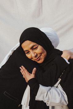 two women hugging each other in front of a white sheet covered wall and one woman is wearing a headscarf