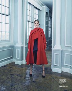 a woman in a red coat and black skirt standing on a tile floor next to columns