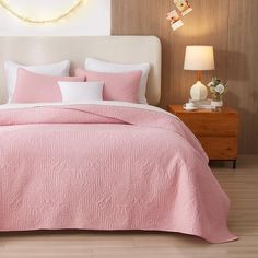 a bed with pink comforter and pillows in a white room next to a night stand