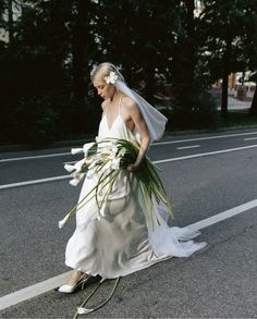 a woman is walking down the street with flowers in her hand and wearing a wedding dress