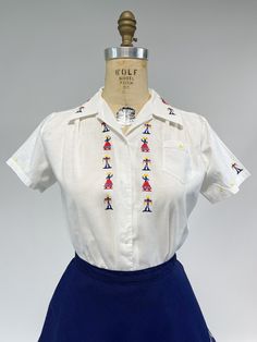 "An utterly adorable vintage blouse from the 1950s! Crisp white linen shirt with button front, small collar and short sleeves. Gentle pleat at each side of bust. Decorated with hand embroidery of little men carrying a milk maids yoke and women in long skirts with hats (or are they carrying fruit?). No labels present, was likely purchased as a souvenir from a vacation taken long ago. Good condition, one button has been replaced. Former museum inventory, has faint hand written number at bottom of Affordable Vintage Tops With Button Cuffs, 50s Shirts For Women, Embroidered Blouses, Embroidered Short Sleeve Daywear Shirt, Embroidered Short Sleeve Day Shirt, Embroidered Short Sleeve Shirt For Daywear, Retro White Embroidered Blouse, Vintage Embroidered Collared Blouse, Vintage Collared Shirt With Floral Embroidery