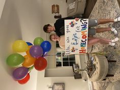 two people holding a sign and balloons in front of a clock on the wall above them