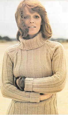 a woman with her arms crossed wearing a turtle neck sweater and gloves, standing in the sand