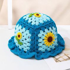 a blue crocheted hat with sunflowers on the front and side, sitting on a table