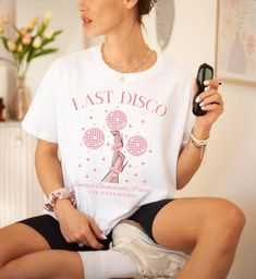 a woman sitting on a chair holding a cell phone and wearing a t - shirt that says last disco