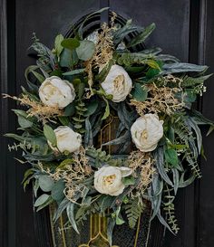 a wreath with white flowers and greenery hanging on a black front door, surrounded by foliage