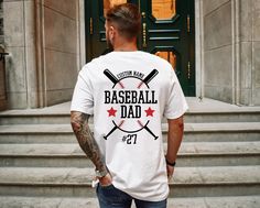 a man wearing a baseball dad t - shirt standing in front of a door with two bats on it