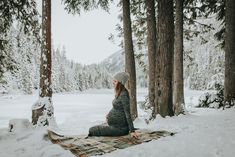 a woman sitting on top of a blanket in the snow next to some pine trees