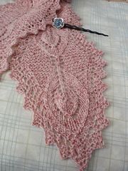 a pink knitted shawl laying on top of a white bed sheet with a black ribbon