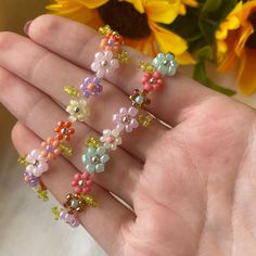 the hand is holding four different colored beaded bracelets with flowers in the background