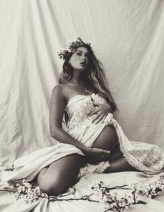 a pregnant woman wearing a flower crown sits on a bed in front of a white backdrop