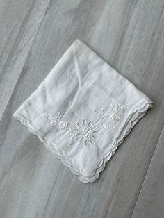 a white napkin with an embroidered flower on it sitting on a wooden floor next to a knife and fork