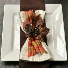 a white plate topped with a napkin covered in fall leaves and pineconis on top of a wooden table