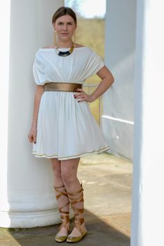 a woman standing next to a pillar wearing sandals and a white dress with gold belt