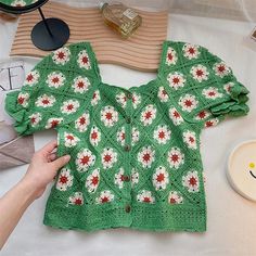 a green sweater with red and white flowers is being crocheted by someone's hand