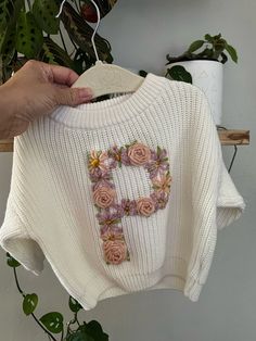 a white sweater with flowers on it being held up by a person's hand