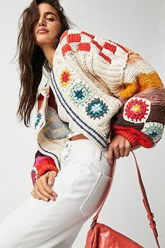 a woman in white jeans and a colorful sweater is holding a red handbag while posing for the camera