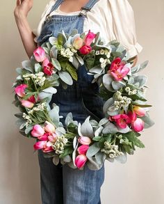 a woman wearing overalls holding a wreath with pink flowers and greenery on it