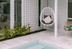 a white hanging chair sitting next to a swimming pool in front of a house on a sunny day