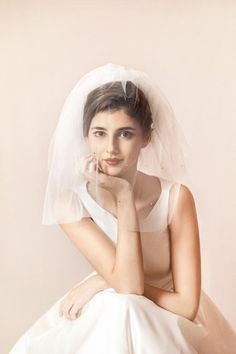 a woman in a white dress and veil sitting on a chair with her hand under her chin