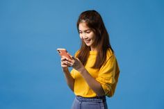 a young woman is looking at her cell phone and smiling while standing against a blue background