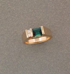 a gold ring with an emerald colored stone and two diamonds on the side, sitting on top of a gray surface