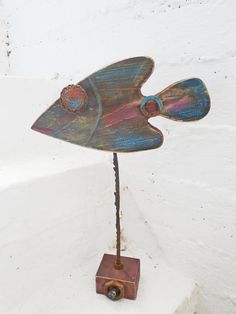 a metal fish sculpture sitting on top of a wooden stand next to a white wall