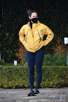 a woman wearing a face mask while standing in front of a hedge with her hands on her hips