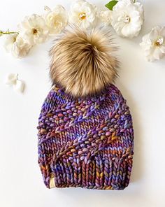 a knitted beanie with a fur pom on top and flowers in the background