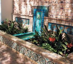 an outdoor fountain with plants and flowers in the center, surrounded by stone tiles on the outside wall