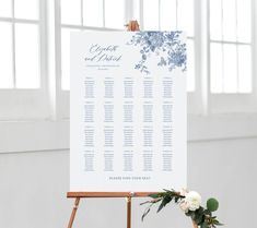 an easel with a seating chart on it next to flowers and greenery in front of windows