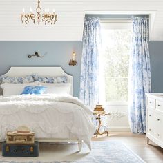 a bedroom with blue walls and white bedding, chandelier, dresser, and window