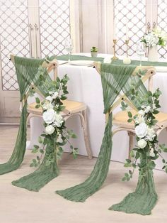 two chairs covered in green cloths with flowers and greenery draped over them at the head table