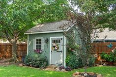 a small shed in the middle of a yard with trees and flowers on it's side