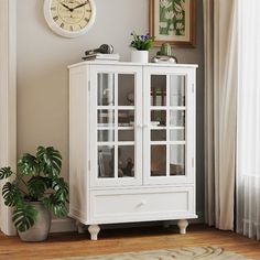 a white cabinet with glass doors in front of a wall clock and potted plant