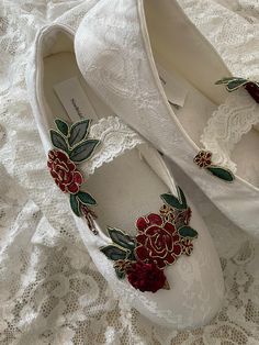 two pairs of white shoes with red flowers on them sitting on a lace covered surface